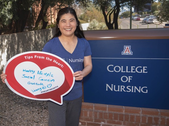 Asian woman with long dark hear wearinc a University of Arizona College of Nursing t-shirt holds up a heart poster outside next to the College of Nursing sign. 