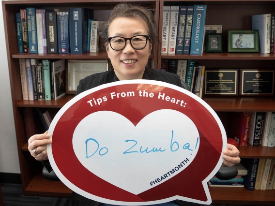 Middle-aged Asian woman with glasses holds up a heart poster in front of a bookcase.