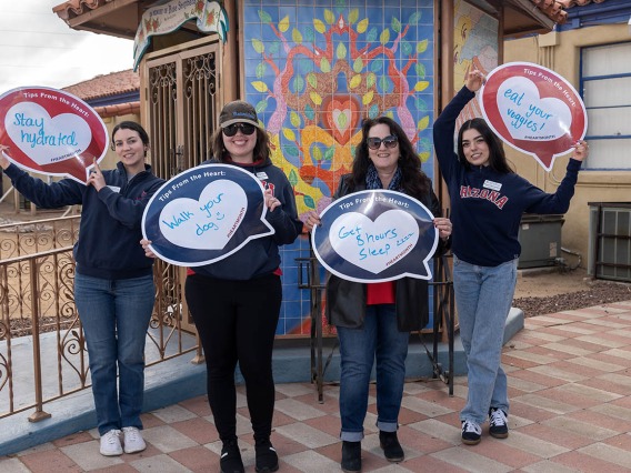 4 woman stand outside in front of a mosaic mural holding heart posters. 