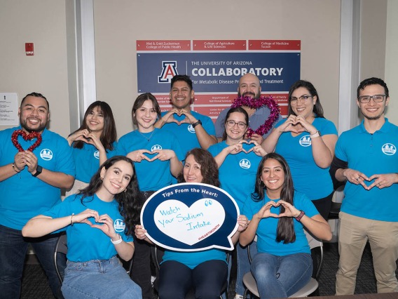 11 smiling people in matching blue shirts make hearts with their hands.