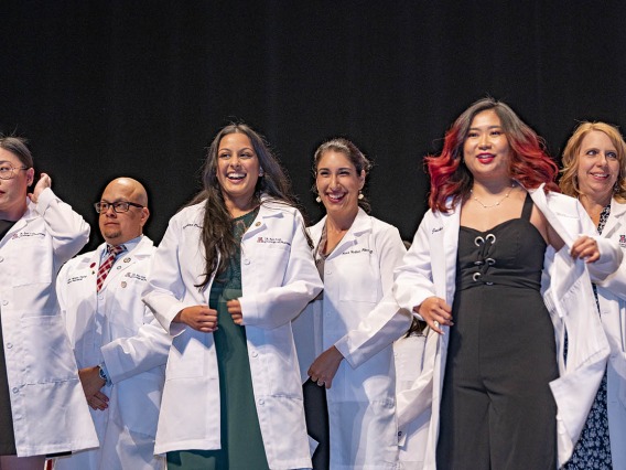 Three female Pharmacy students are presented their white coats by faculty members standing behind them on a stage. 