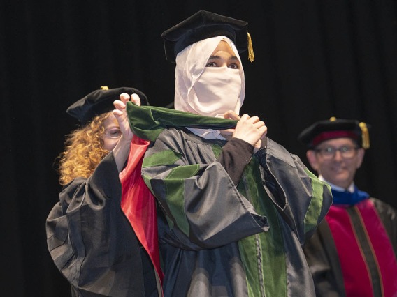 A young woman wearing graduation regalia and a head and face covering is presented with a sash for graduation.