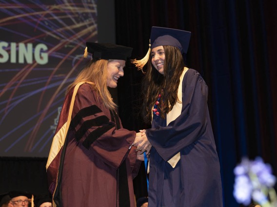 A young woman with long brown hair shakes hands with a female professor on stage during convocation.