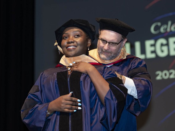 A young woman wearing graduation regalia smiles as she receives her hood by a male professor during convocation.