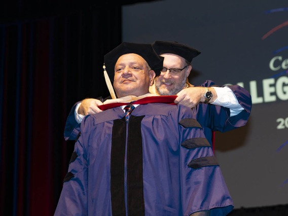 A man dressed in graduation regalia receives his hood from a male professor during graduation.