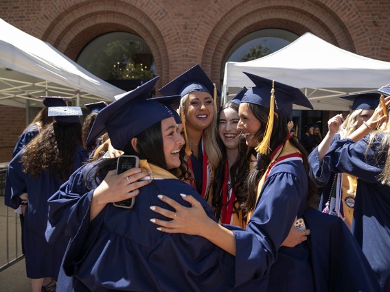 A group of young women wearing cap and gown celebrate together in a group hug after their convocation. 