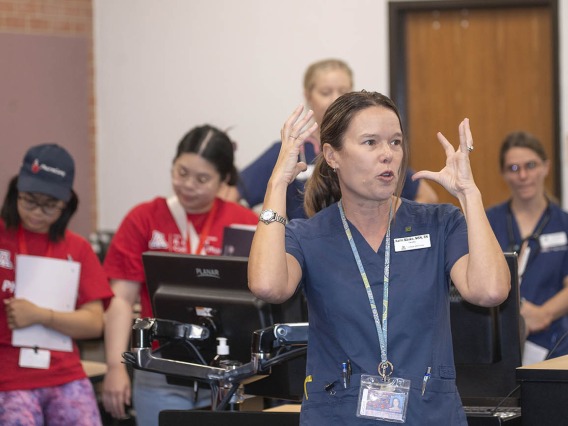 A middle-aged light skinned woman in scrubs motions with her hands as she is talking to studetns.