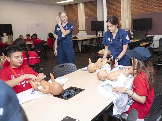 A nursing student in blue scrubs shows several young students how to change a diaper on a baby manikin.