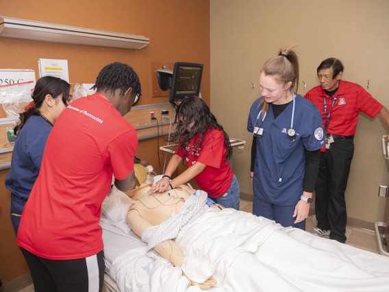 Two kids in practice CPR on a manikin in a clinical setting while nursing students and a professor observe.