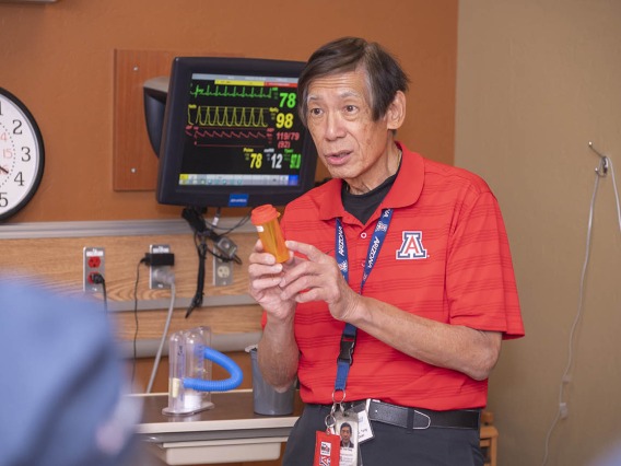 An older Asian man stands in a hospital room setting holding a prescription bottle while talking.