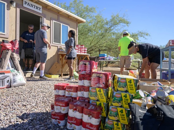Stacks of canned goods sit on the ground outside of a small building with a sign that reads, "Pantry", while a few people outside the building are moving items into it.