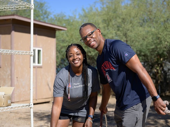 A young black woman and man stand next to each other smiling as they pause from volunteer work outside a small building. 