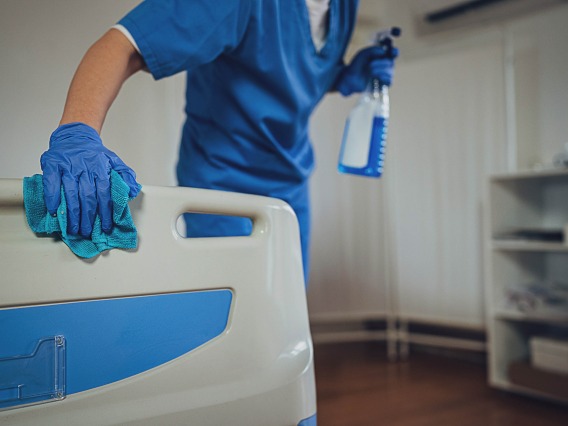 A nurse in blue scrubs wipes down a hospital bed with one hand while holding a spray bottle in the other.