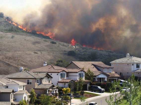 A wildfire burns a vegetated area behind a row of houses; flames and smoke are visible.