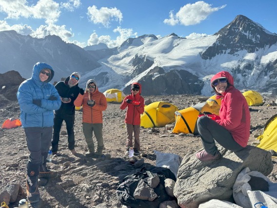 Snow-covered mountains are in the background as five men and women, all dressed in parkas, hold bowls of food.