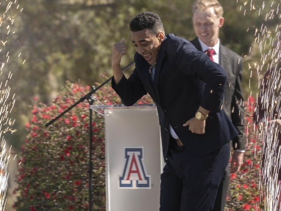 A young adult male wearing a dark suite does a dance on a stage with sparklers going off around him. A University of Arizona logo is behind him. 