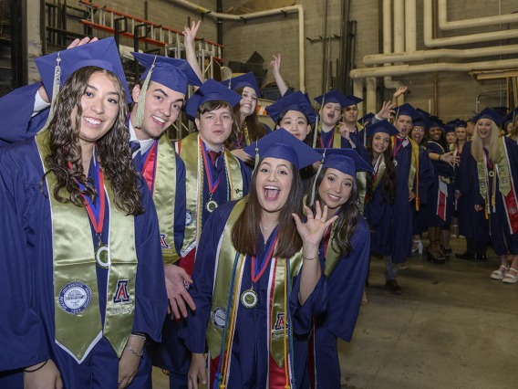A large group of graduates in blue caps and gowns smile and cheer in a line backstage before their graduation ceremony. 