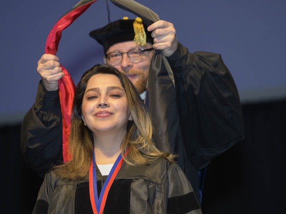 A young woman with light brown hair wearning a graduation gown smiles as a professor places a sash over her shoulders. 