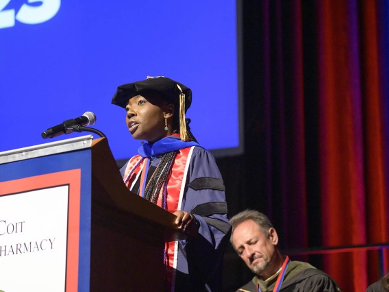 A young Black woman in graduation cap and gown talks into a microphone at a podium. 
