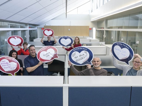 7 people standing up in cubicle area of office hold heart posters.