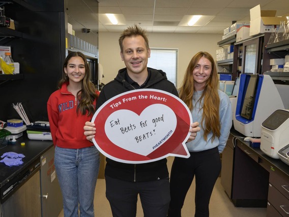 White man in dark hoody stands in lab holding a heart poster with two female students standing slightly behand him.