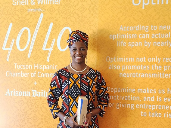 Dark-skinned woman wearing a head wrap and patterened dress stands in front of a 40 Under 40 banner holding an award and smiling. 