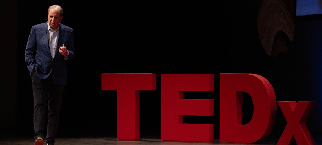 Dr. Michael Dake walks across a stage in front of a large, red TEDx sign.