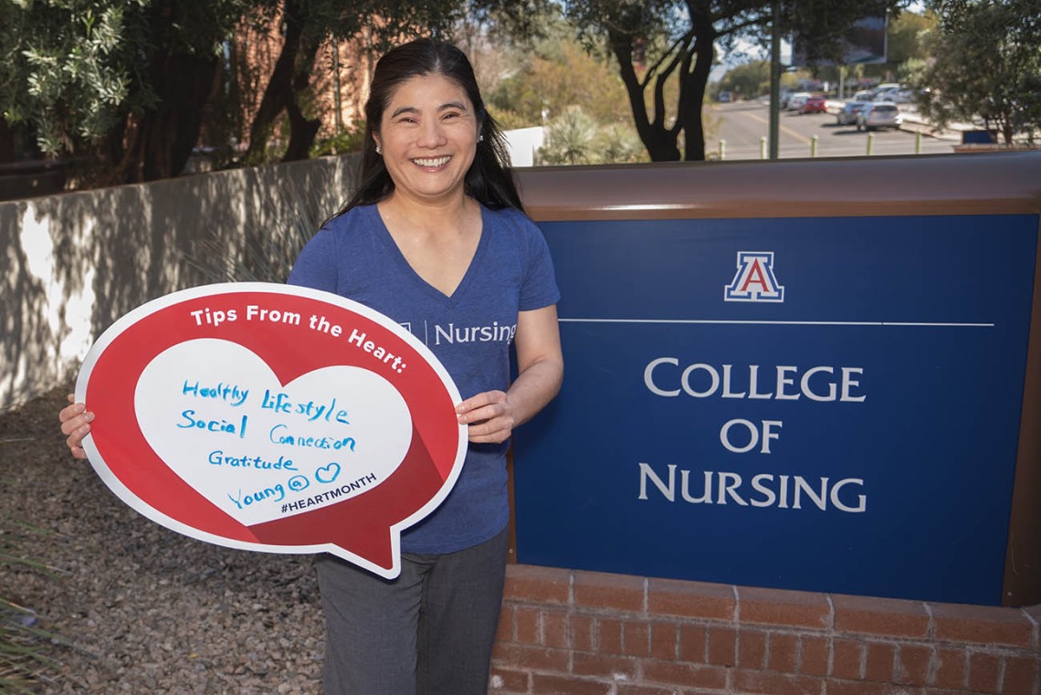 Asian woman with long dark hear wearinc a University of Arizona College of Nursing t-shirt holds up a heart poster outside next to the College of Nursing sign. 
