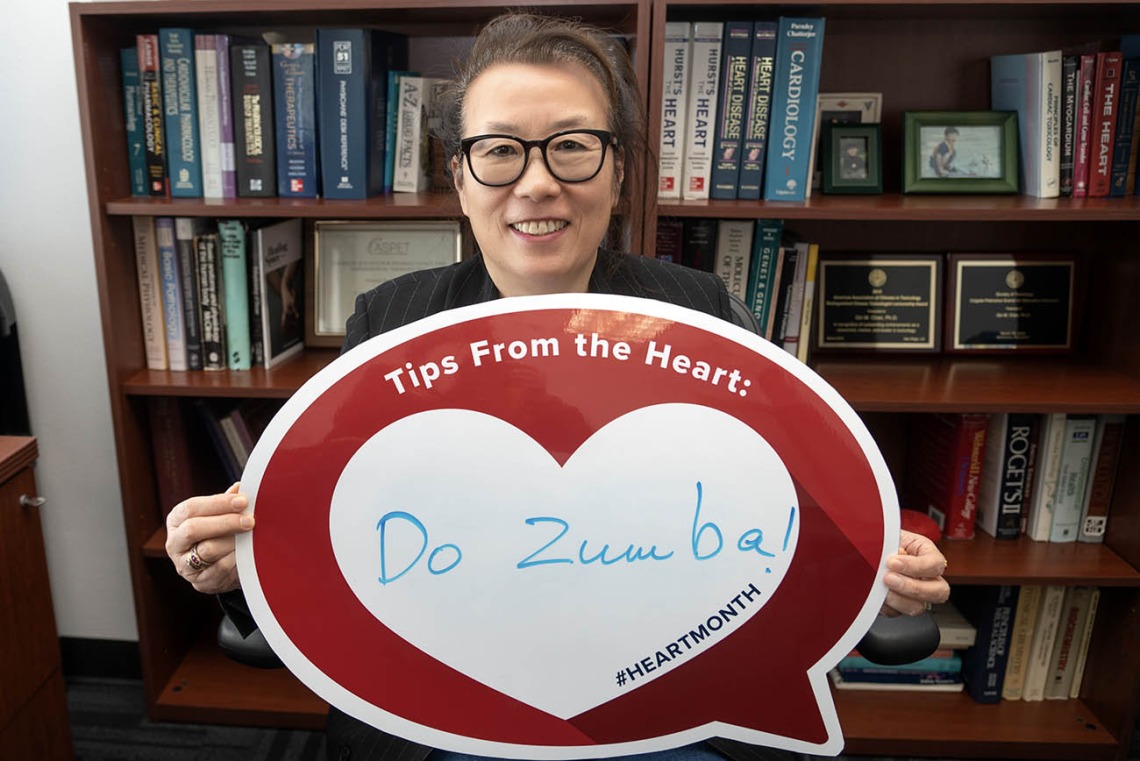 Middle-aged Asian woman with glasses holds up a heart poster in front of a bookcase.