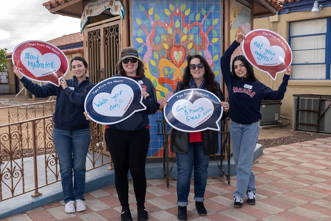 4 woman stand outside in front of a mosaic mural holding heart posters. 