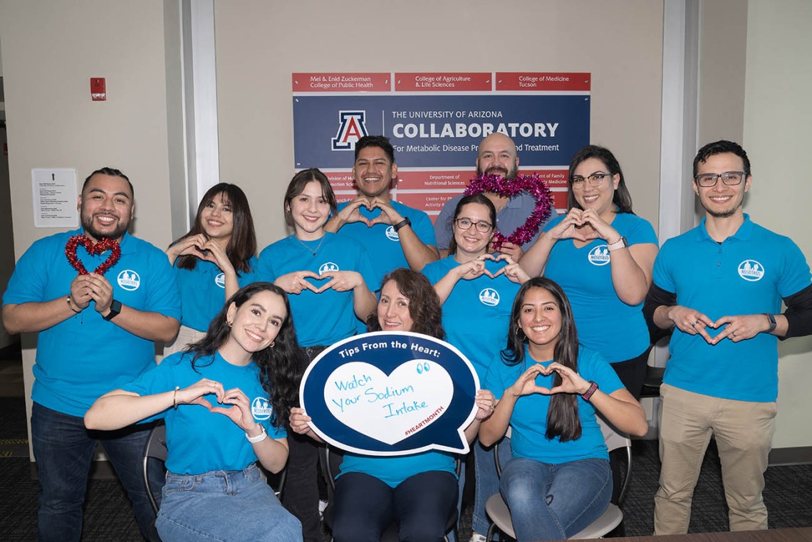 11 smiling people in matching blue shirts make hearts with their hands.
