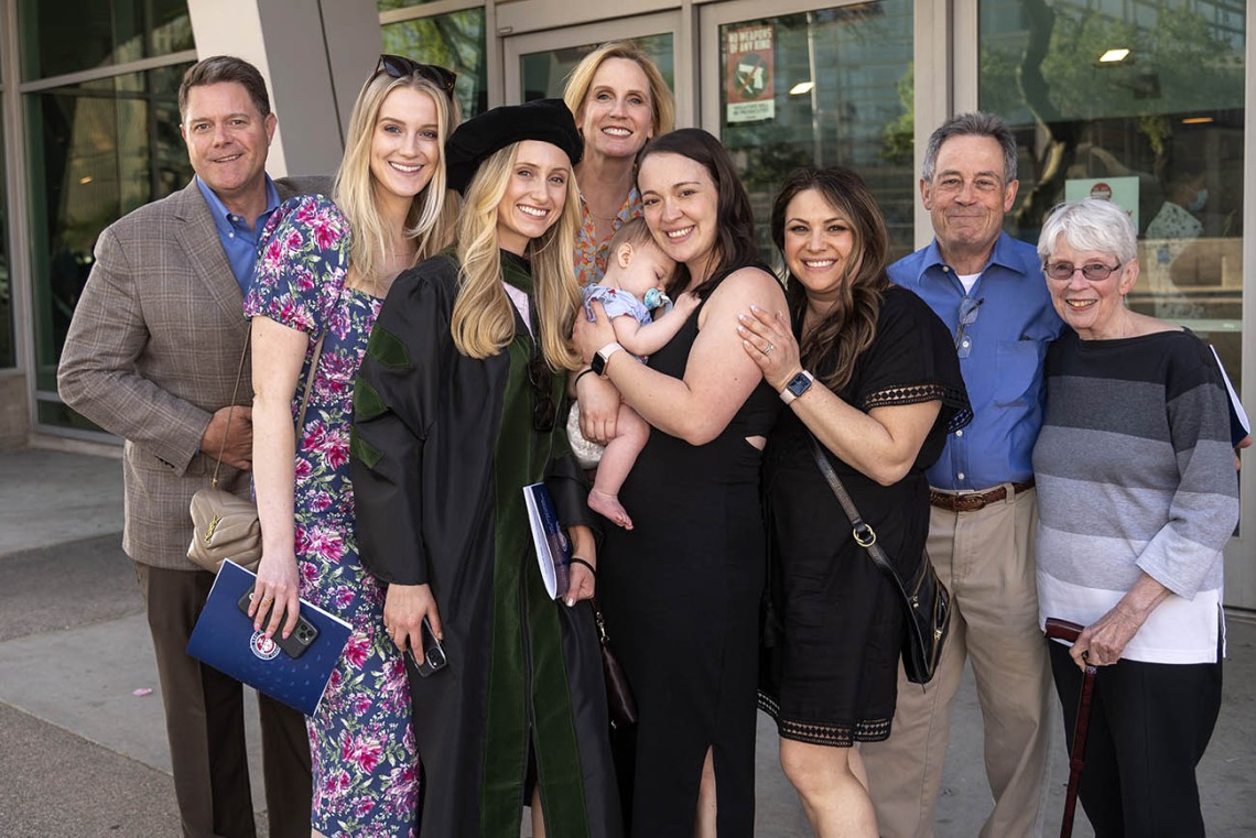 A light-skinned young blonde woman in graduation regalia stands with several members of her family, all smiling. One woman is holding a sleeping baby. 