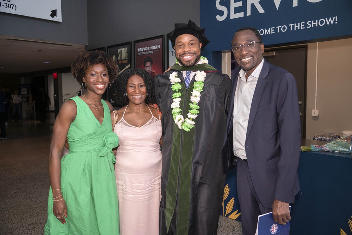 A tall dark-skinned man in graduation regalia stands with his parents and girlfriend, all smiling. 