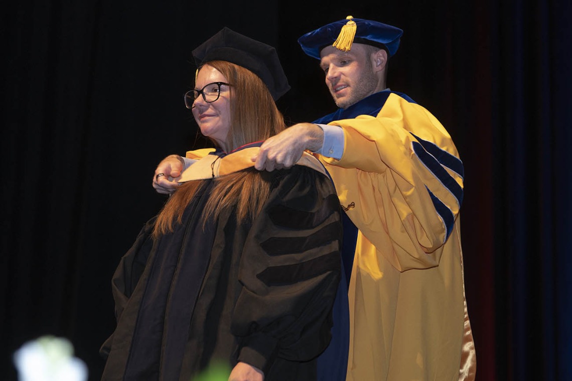 A young woman with red hair and wearing graduation regalia receives her hood by a male professor during convocation.