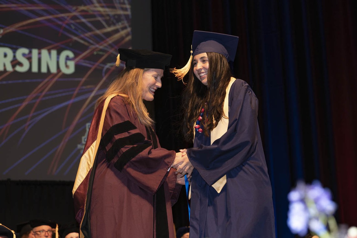 A young woman with long brown hair shakes hands with a female professor on stage during convocation.