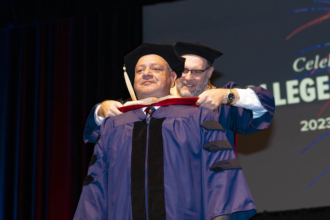 A man dressed in graduation regalia receives his hood from a male professor during graduation.