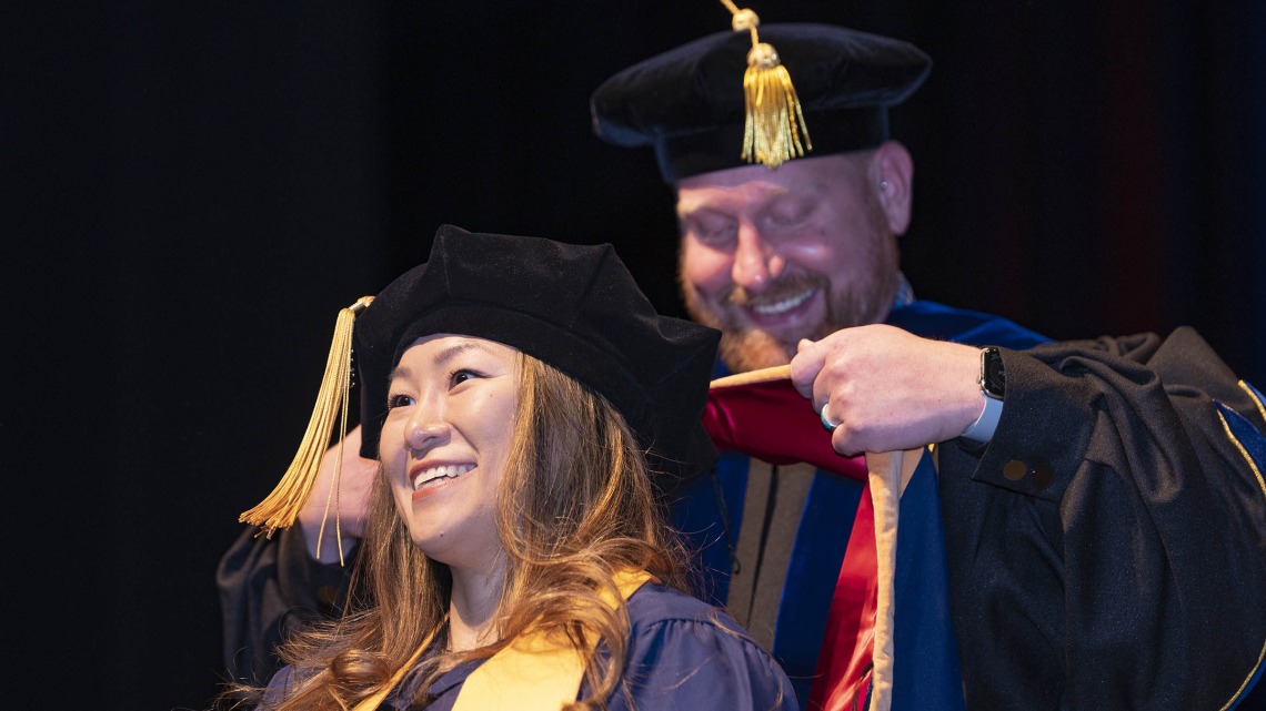 Woman with brown hair wearing graduation regalia smiles while waiting to be hooded by a male professor.