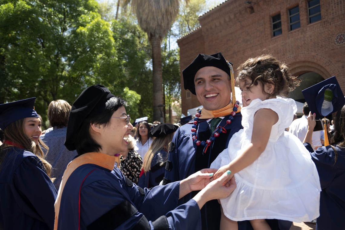 A man dressed in graduation regalia holds a toddler girl wearing a white dress. Another woman wearing graduation cap and gown reaches out to hold the toddler’s hand while smiling in celebration. 
