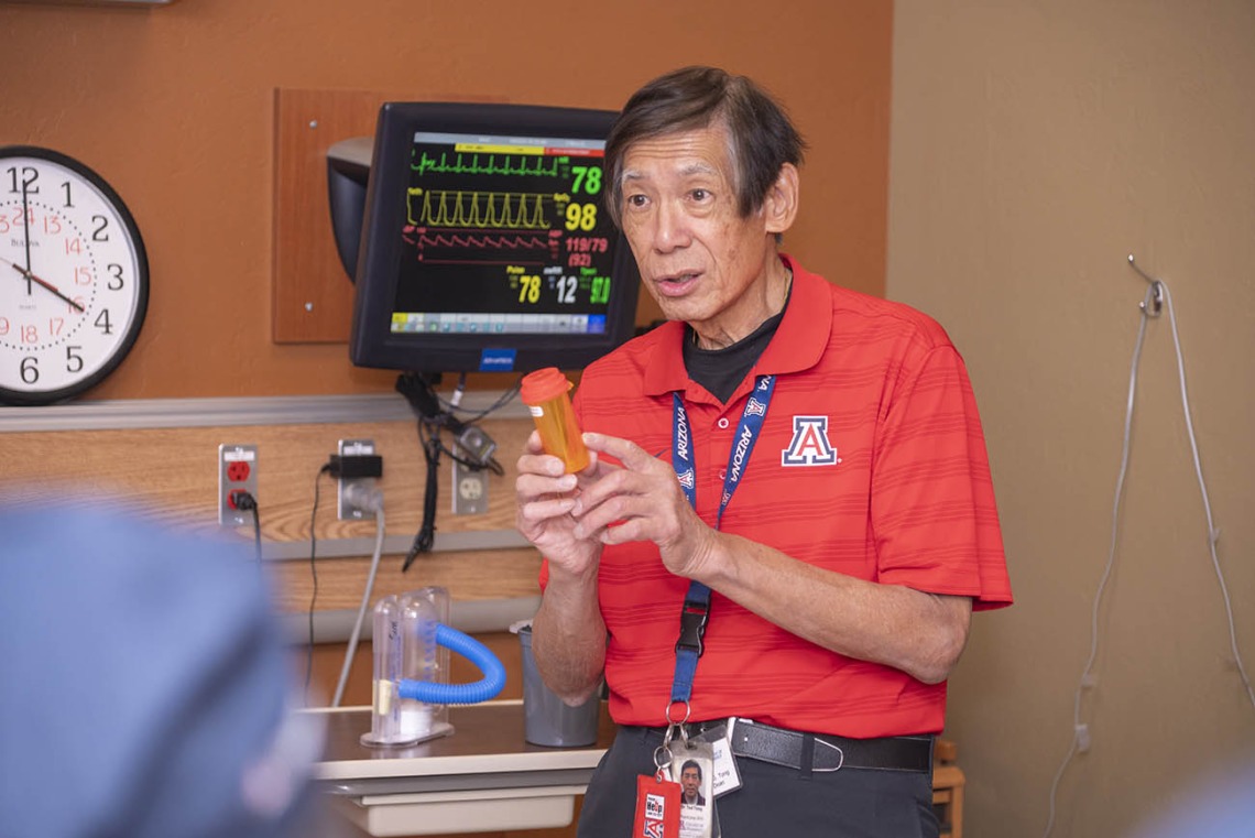An older Asian man stands in a hospital room setting holding a prescription bottle while talking.