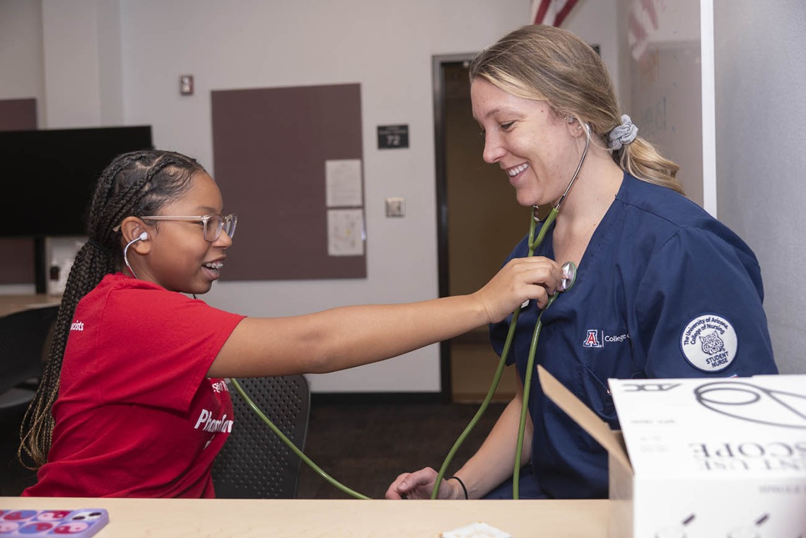 A young dark-skinned girl uses a stethoscope to listen to the heartbeat of a nursing student. 