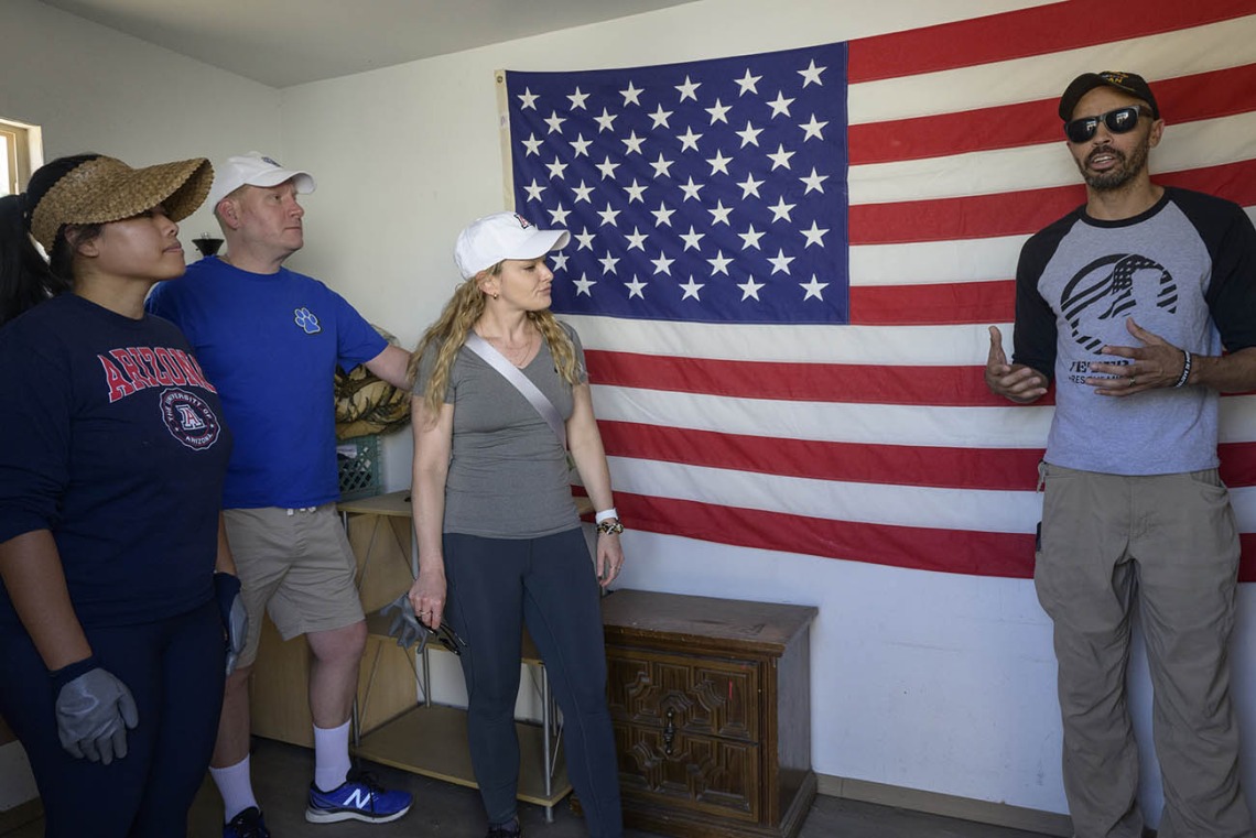 A tall man in sunglasses stands in front of an American flag talking to three volunteers from the UArizona nursing program.