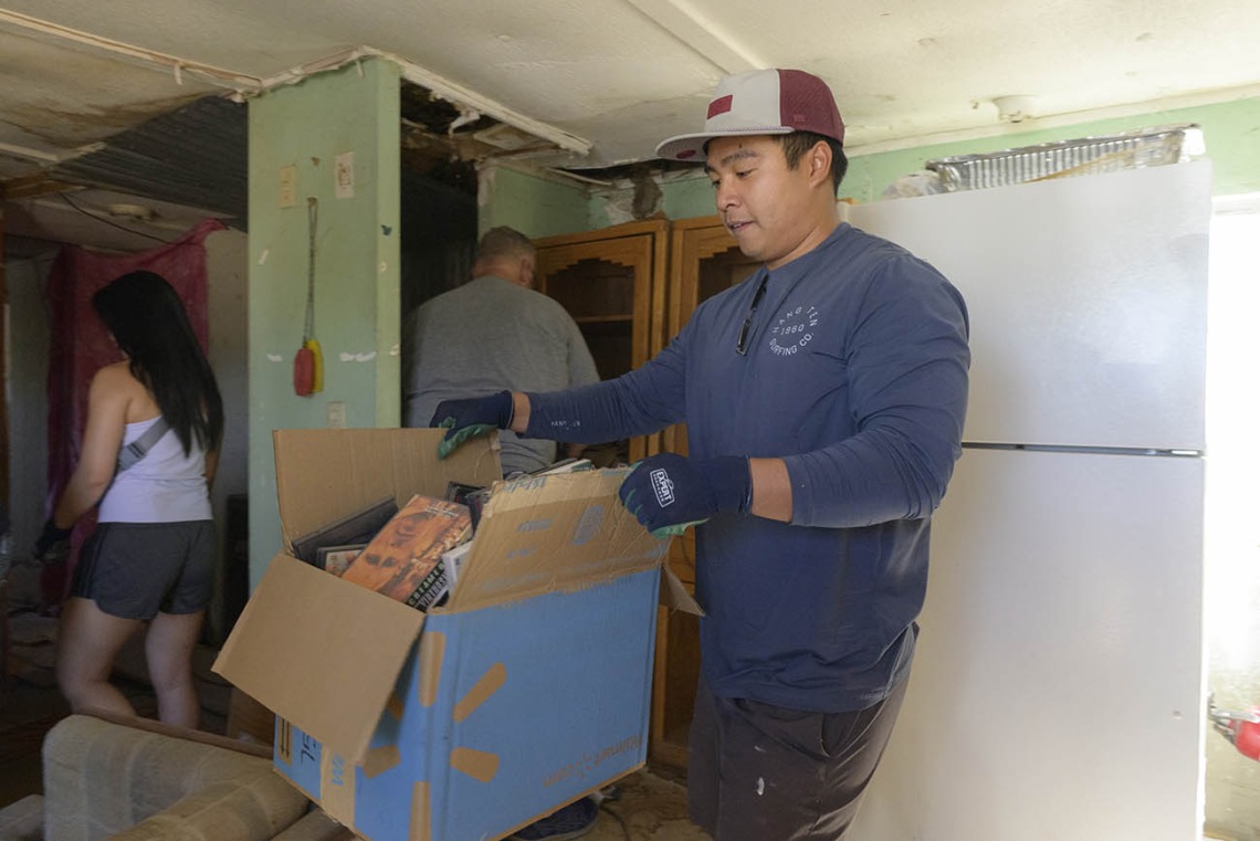 A young man wearing a baseball hat and gloves carries a large box through a rough-looking room. 