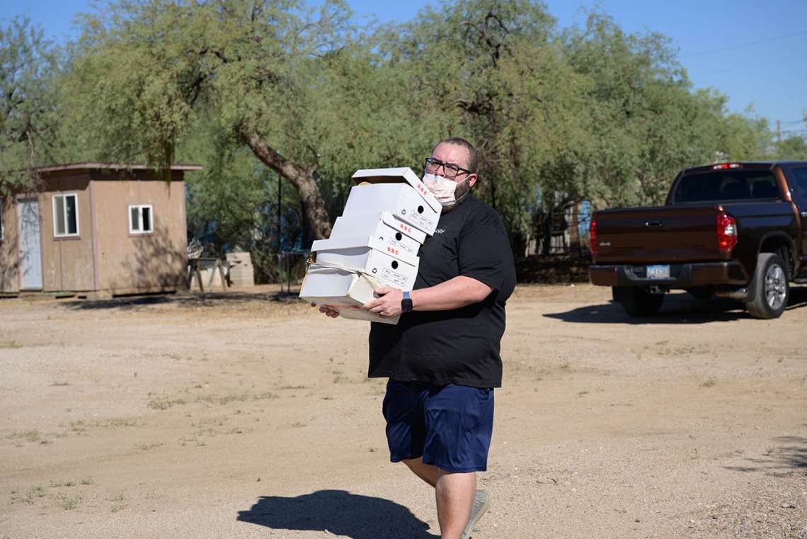 A man carries a stack of shoe boxes across a dirt parking lot. 