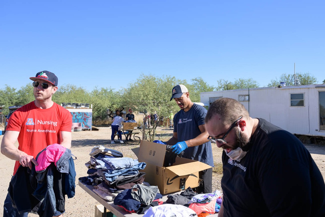 Three young men, all Doctor of Nursing Practice residents, sort donated clothes at a table covered in clothing in a dirt lot. 