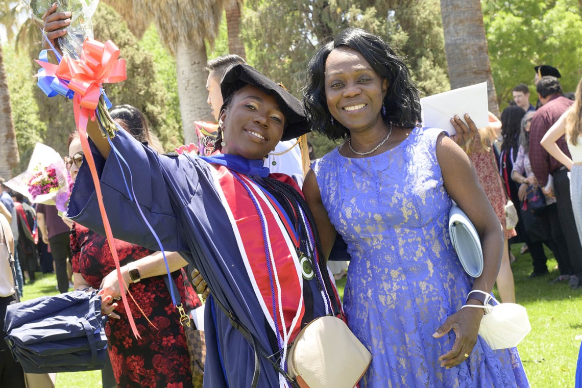 Two young Black women, one in a dress and the other in a graduation cap and gown, side-hug and smile.