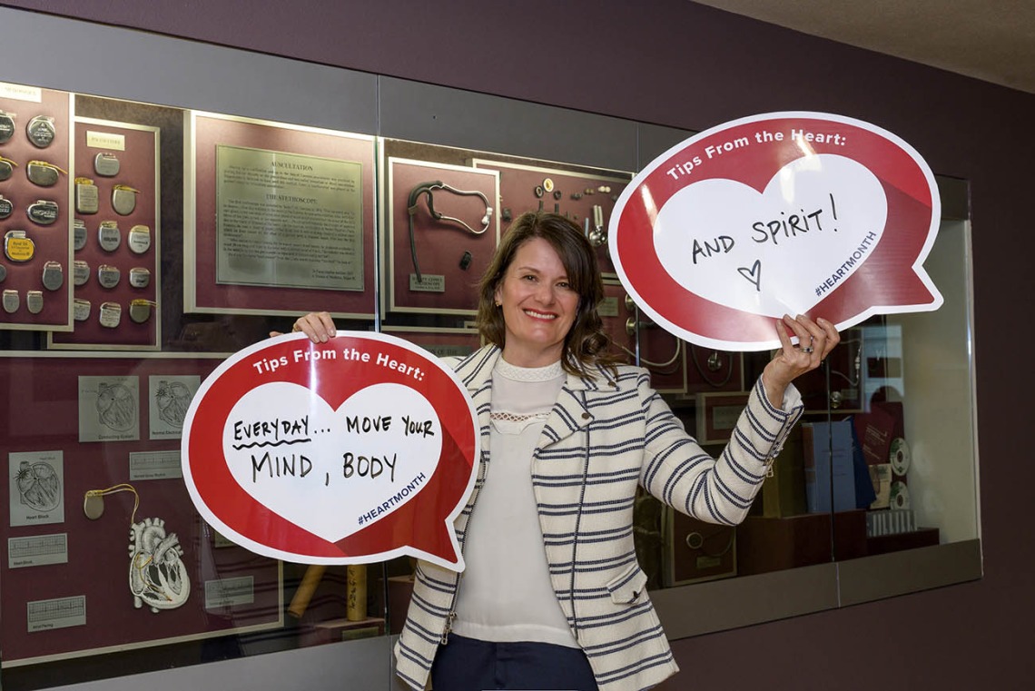White woman with brown hair and and striped jacket holds up two heart posters in front of a display cabinet