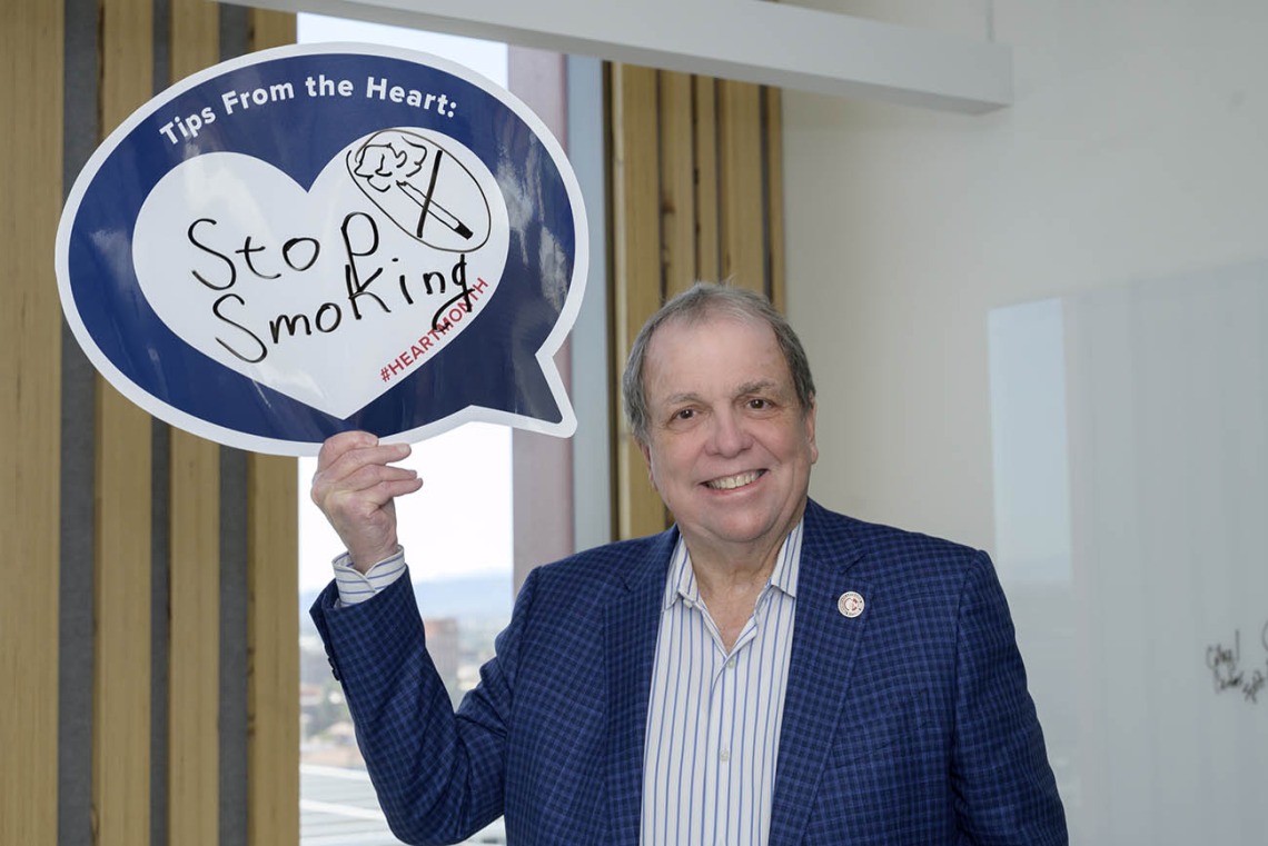 Older white man in blue sport coat holds up sign that says Stop Smoking!