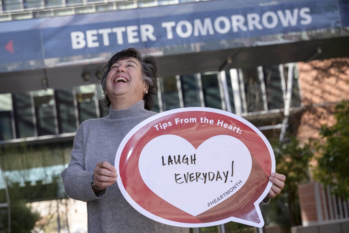 Middle-aged woman with short dark hair laughs as she holds a heart poster that says Laugh everyday!