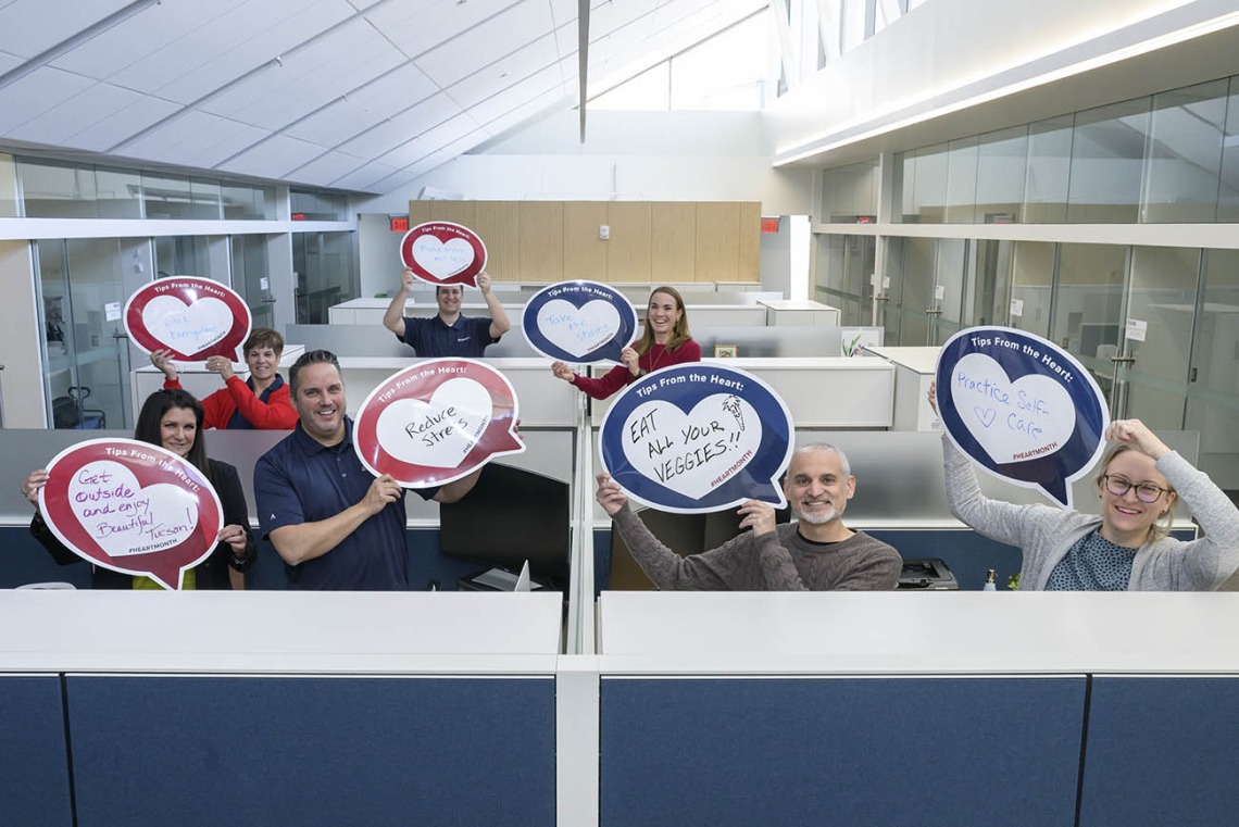 7 people standing up in cubicle area of office hold heart posters.