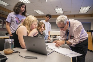 An older male doctor wearing glasses works with college-aged students at a table.